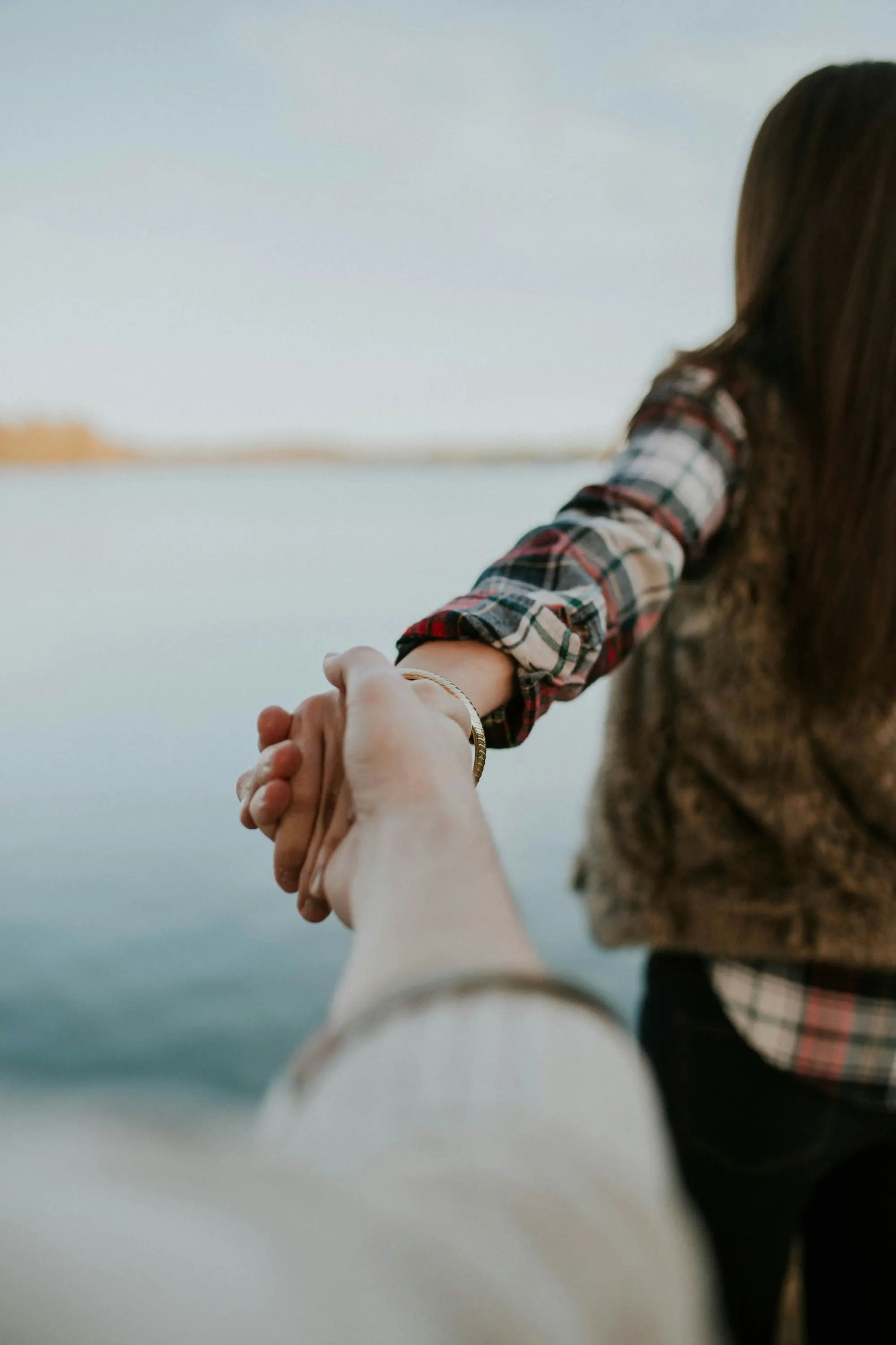 A person with long hair and plaid shirt leads another, holding hands near a calm body of water, evoking a sense of adventure.