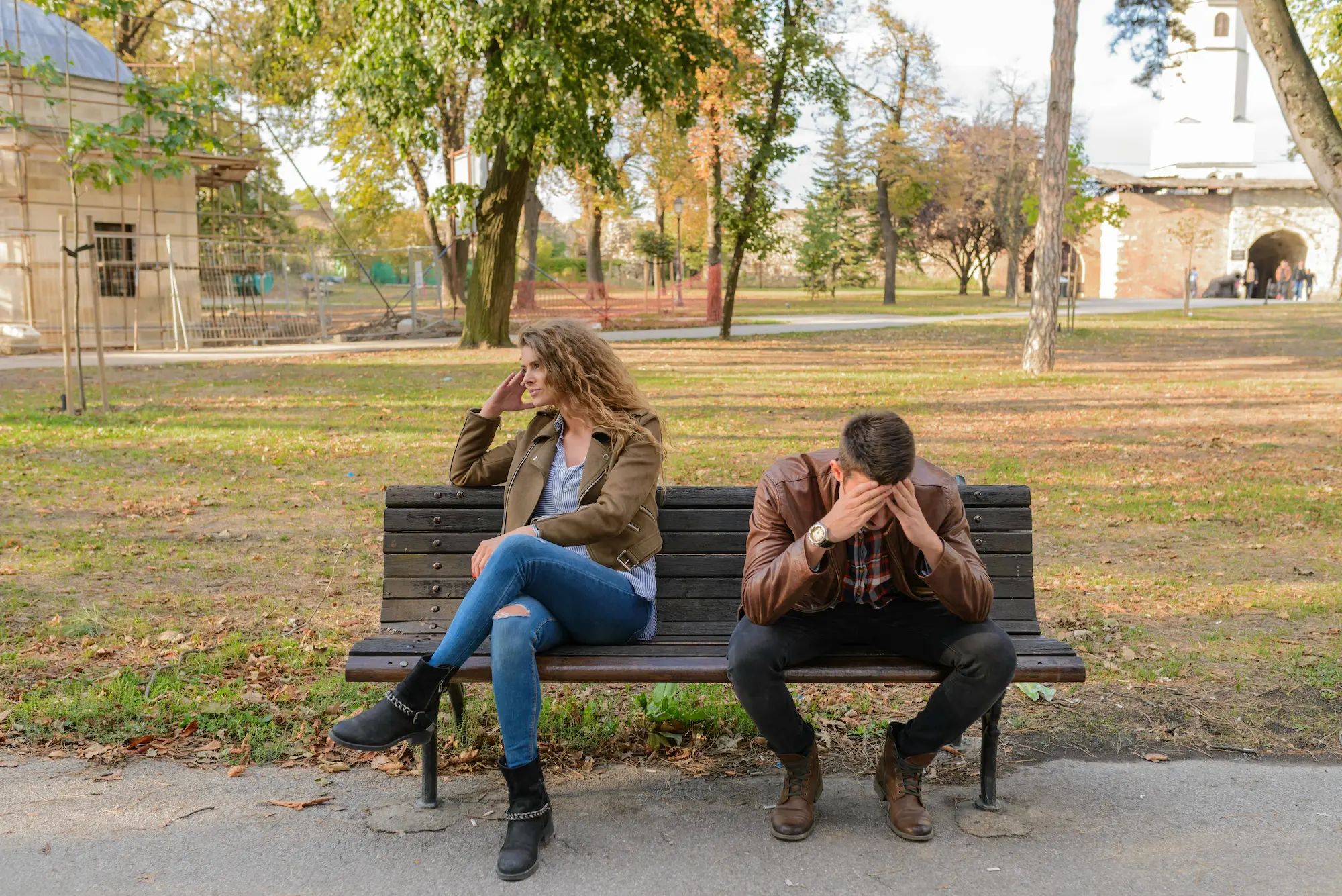 Two people sit on a park bench, looking upset and facing away from each other. Trees and a path are in the background.