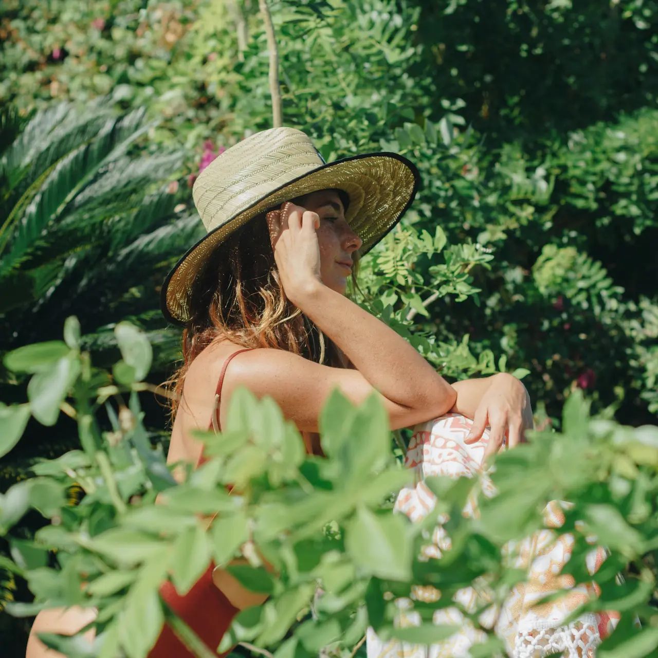 Woman in a straw hat sits in lush greenery, enjoying a sunny day.