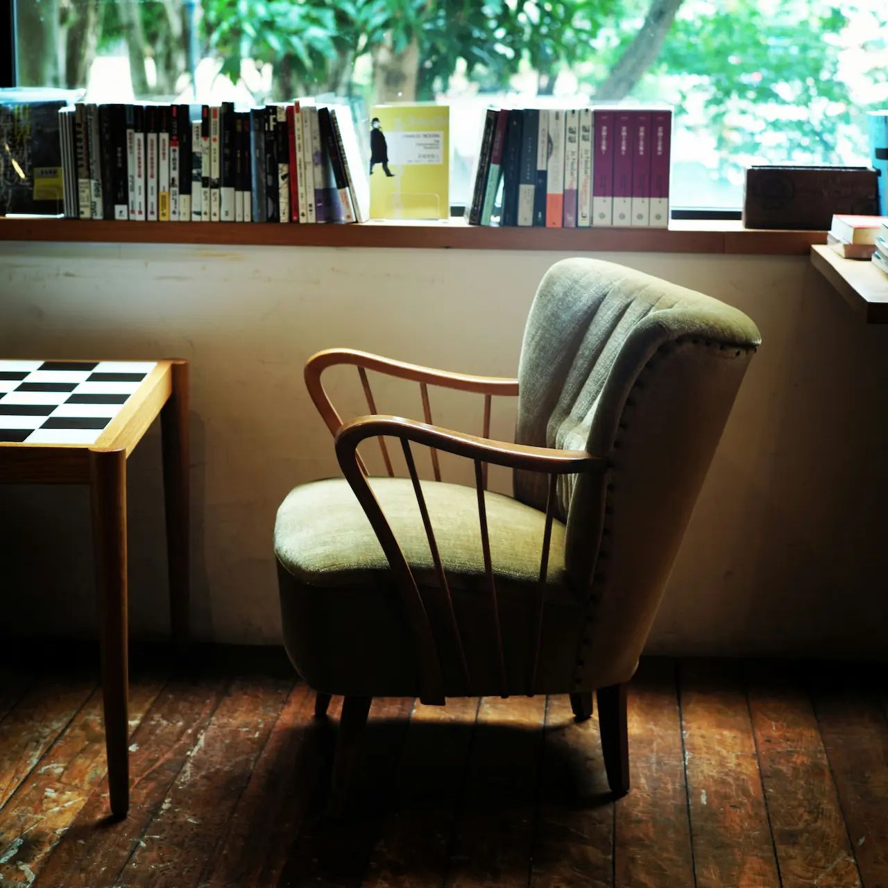 Cozy reading nook with an armchair, wooden floor, bookshelf, and a chessboard table by a window.