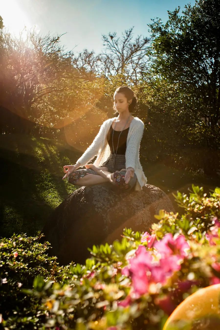 Woman meditating on a rock in a sunlit garden, surrounded by blooming flowers and greenery.