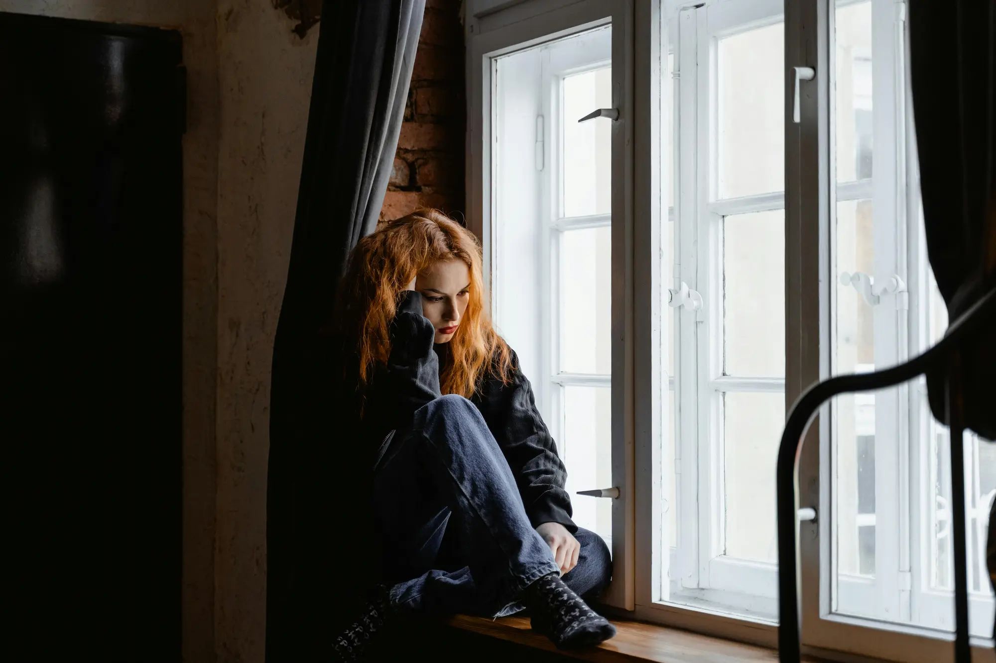Red-haired woman sitting on a windowsill, looking contemplative, with natural light streaming through a white-framed window.