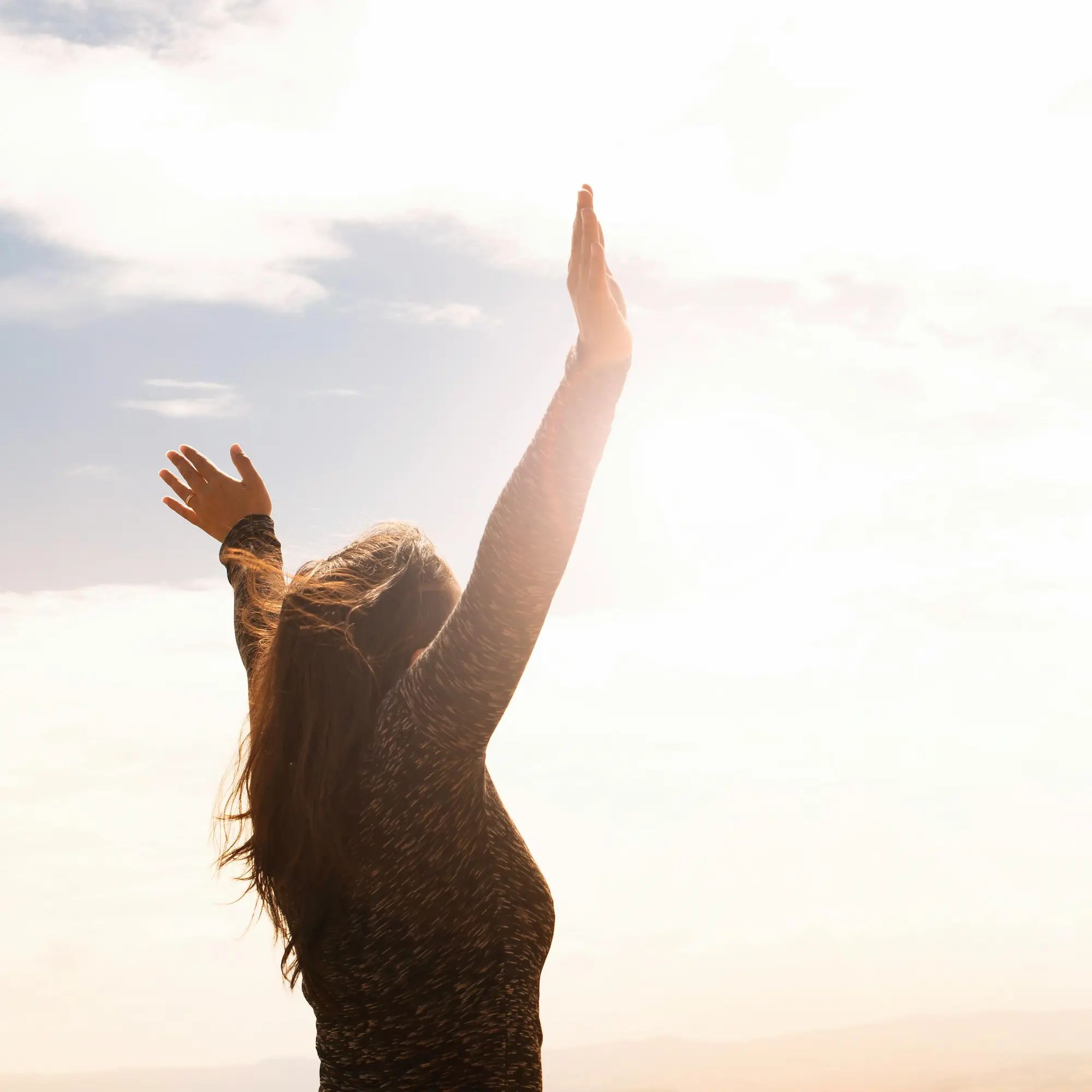 Person with raised arms in sunlight, wearing a long-sleeve shirt, expressing freedom or joy.