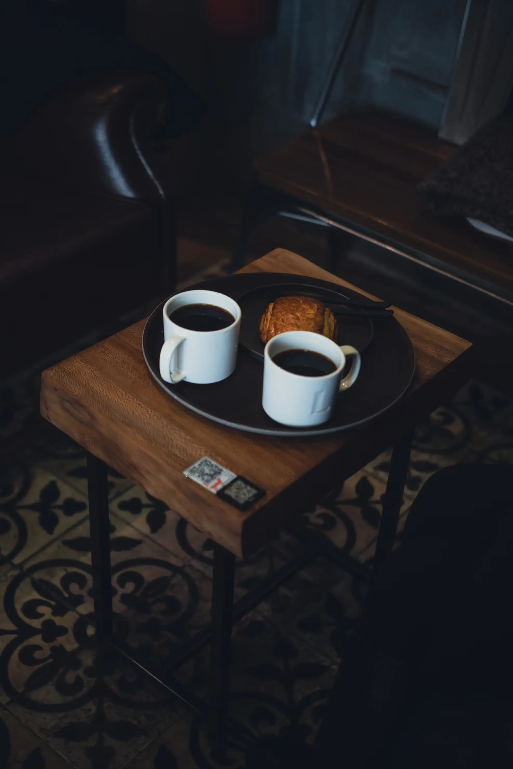 Two coffee mugs and a pastry on a tray atop a wooden side table with a tiled floor pattern.